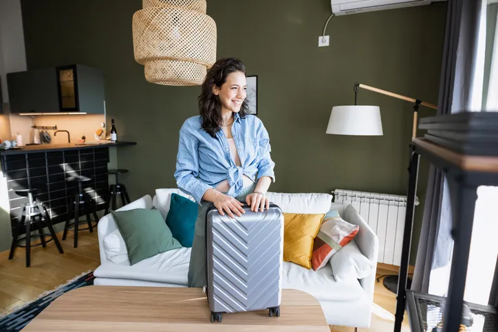 Photo of a young woman at home, standing with suitcase and waiting for taxi