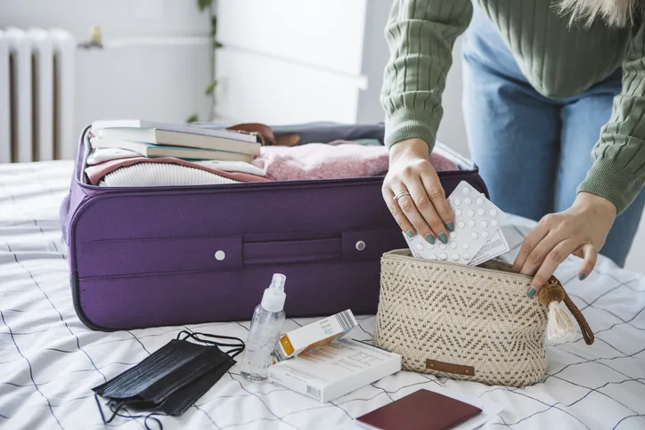 Woman preparing travel suitcase on bed at home. She is packing medicine and disinfection stuff for travel.