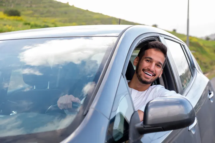 A cheerful young man with a beard, wearing a traditional Middle-Eastern outfit, smiles confidently while seated in his car