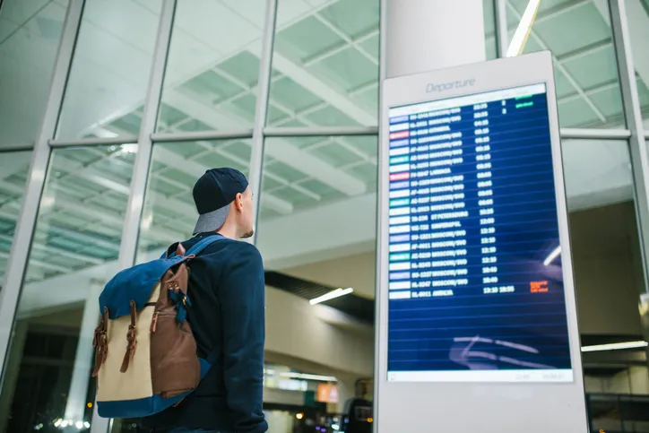 A young male traveler with a backpack in casual style looks at the information board at the airport. Getting information about the flight.